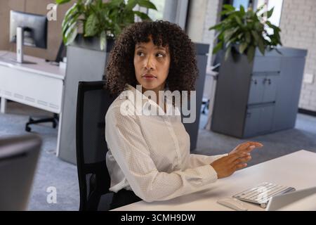 African american woman sitting at office desk looking over shoulder next to white keyboard, mouse Stock Photo