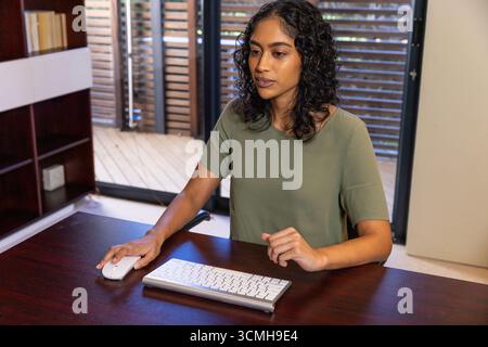 Indian woman typing on wireless keyboard with mouse at dark wood desk in home office Stock Photo