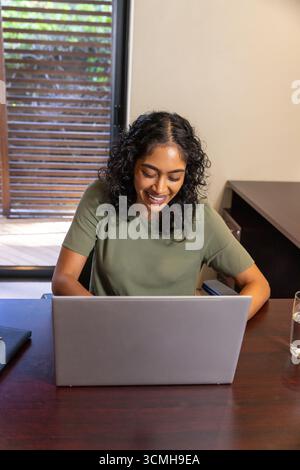 Indian woman smiling while typing on laptop at wooden desk with notebook and water glass Stock Photo