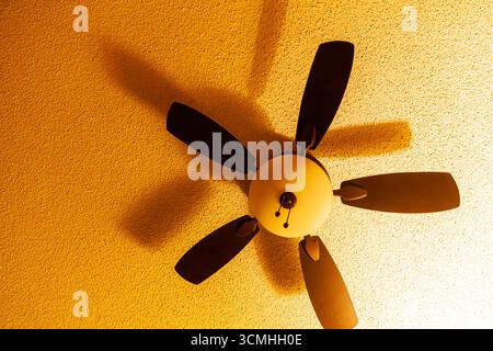 Ceiling fan casting a rotating shadow on the ceiling surface with soft yellow light, close up Stock Photo