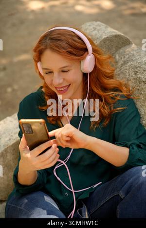 Cheerful woman enjoying music using pink headphones and browsing smartphone while relaxing in park Stock Photo