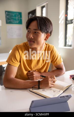 Teenage asian male student writing geometric in notebook in classroom, tablet nearby Stock Photo