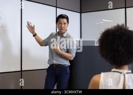 Diverse coworkers presenting on whiteboard at lectern in meeting room with marker and laptop Stock Photo