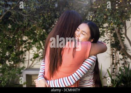 Diverse female friends hugging in private backyard garden with climbing ivy and bench Stock Photo