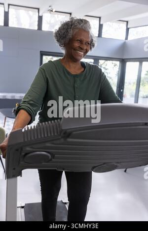 Senior african american woman in sportswear using treadmill in gym with seating area, windows Stock Photo
