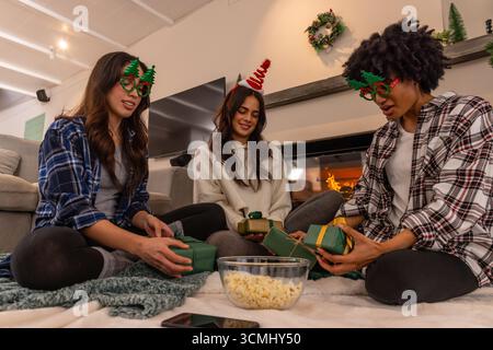 Diverse female friends in headbands and glasses holding gifts with popcorn by cozy fireplace Stock Photo