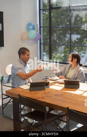Diverse coworkers clinking mugs and sharing coffee break with tablets in room, balloons Stock Photo