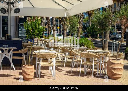 An outdoor cafe with wicker furniture, white tables and greenery under a canopy with fans in a sunny Mediterranean town with palm trees. Stock Photo