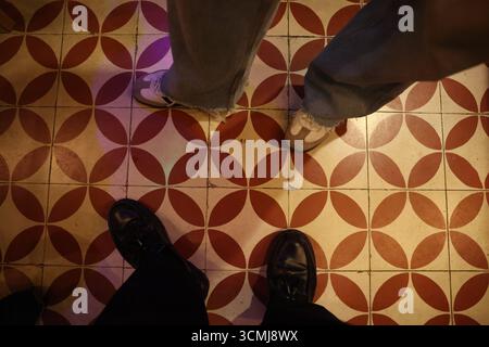 Overhead shot of feet in casual shoes on a red and beige patterned tile floor.  Ideal for travel, fashion, or lifestyle advertising. Stock Photo