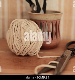 A set of vintage sewing scissors sit on a rustic wood desk with a ball of cotton cord. In the background, a neutral tapestry hangs against the wall. Stock Photo