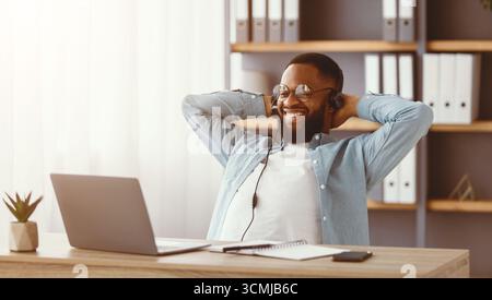 Happy young black bearded man in glasses and headphones rejoices in successful deal, relax and looks at laptop Stock Photo