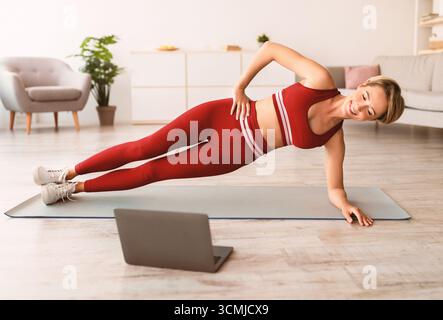 Smiling woman doing side plank on mat with pc Stock Photo