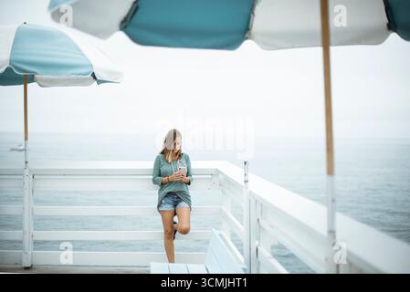 Woman in denim shorts leaning against wooden railings by the ocean looking at her phone, Malibu, California, USA Stock Photo