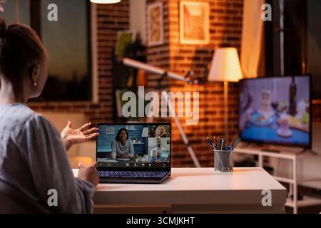 Self employed woman joins video conference with diverse professionals, working remotely in brick wall apartment. Female entrepreneur doing business presentation during virtual meeting with clients. Stock Photo