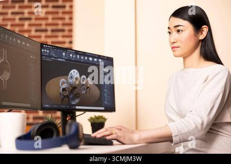 Young female engineer developing industrial 3D models with gears and clamps, using cad software on computer. Remote asian woman building digital prototypes from modern home office environment. Stock Photo