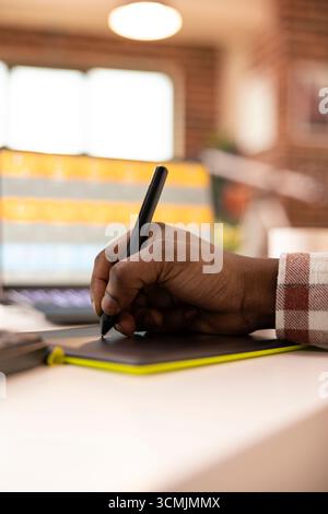 Close up of african american hand using stylus and graphic tablet to retouch images on laptop. Black freelancer utilizing efficient digital photo editing setup in creative home workspace. Stock Photo