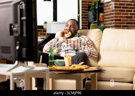 African american male individual relaxing at home during weekend, eating juicy burger and watching TV. Young black man having casual afternoon in apartment with fast food and entertainment on screen. Stock Photo
