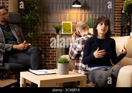 Marriage therapy session with African American psychologist observing as caucasian couple discusses relationship struggles. Husband and wife sit back to back, sharing feelings during counseling. Stock Photo