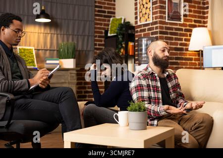 Professional family psychologist sits in chair, listening to clients seated back to back during marriage counseling. African American therapist analyzing relationship issues during couple therapy. Stock Photo