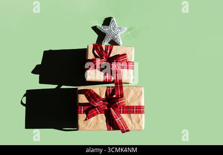 Stack of holiday gift boxes wrapped in craft paper with red plaid ribbon and glitter star topper on green. Christmas present and celebration concept. Stock Photo