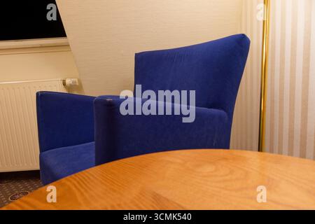 A soft blue velvet armchair stands by a round wooden coffee table. A radiator sits behind, and the roof windows lean inward, giving a top floor feelin Stock Photo
