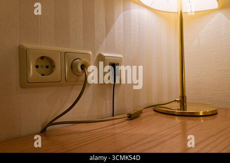 A set of three outlets sits on the wall, two standard sockets and one data style port. A desk lamp is plugged in, and several leads run toward a wall Stock Photo