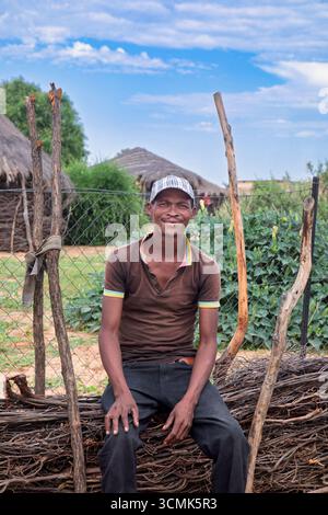 village african man single poverty ,sitting on the fence, wearing a cap, rural living thatched roof mud houses Stock Photo