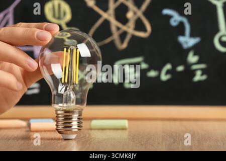 Female hand with light bulb and chalk on wooden table against chalkboard with science drawings, closeup. Inventor concept Stock Photo