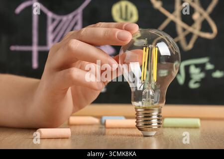 Female hand with light bulb and chalk on wooden table against chalkboard with science drawings, closeup. Inventor concept Stock Photo