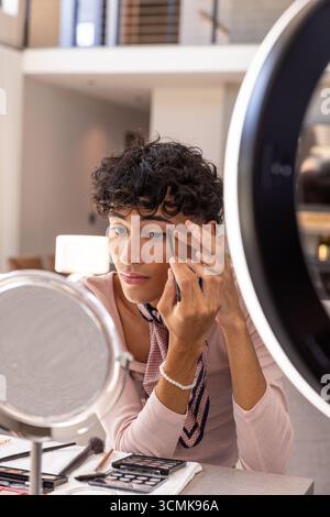 Man applying eyeliner while using mirror under ring light with palettes in living area Stock Photo