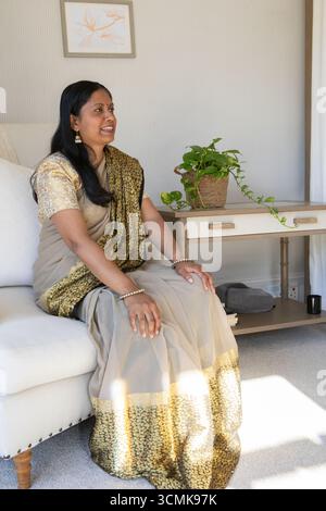 Mature indian woman wearing saree sitting on cream sofa beside table with potted plant Stock Photo