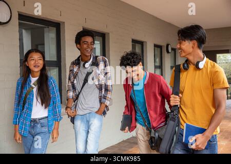 Diverse friends walking and laughing along school walkway carrying backpacks headphones and devices Stock Photo