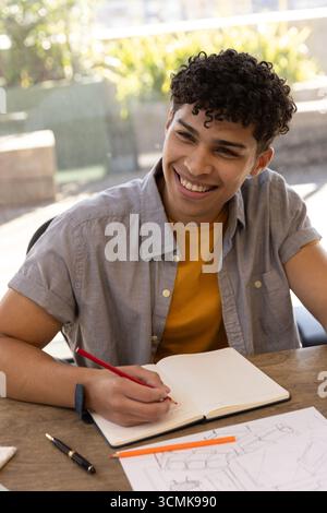 Man smiling while writing in notebook with red pencil and sketch paper on wooden table Stock Photo