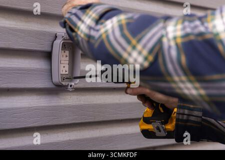Electrician working on installing an outdoor electrical outlet on vinyl siding using a cordless drill Stock Photo