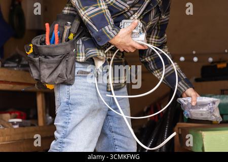 Electrician working on electrical system installation, holding cables and components, wearing tool belt with various tools Stock Photo