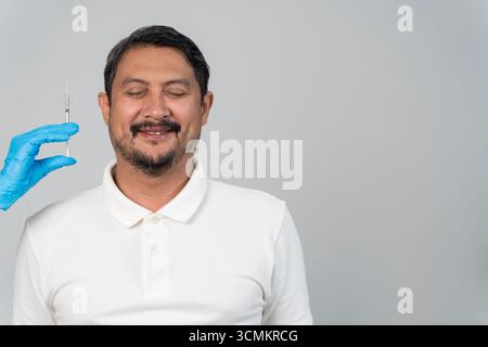 Smiling middle aged man with eyes closed preparing for cosmetic botox injection, doctor wearing gloves holding syringe, concept of beauty care, wrinkl Stock Photo