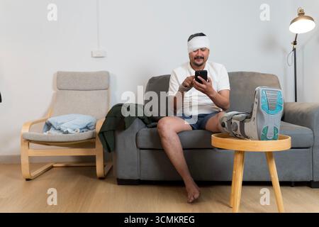 Middle aged man sitting on sofa at home with head bandage and orthopedic leg boot cast, using smartphone while resting, symbolizing injury recovery, h Stock Photo