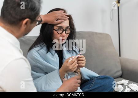 Unwell woman sitting on sofa checking thermometer while man touches her forehead, showing fever symptoms, illness, flu, and home healthcare support, c Stock Photo