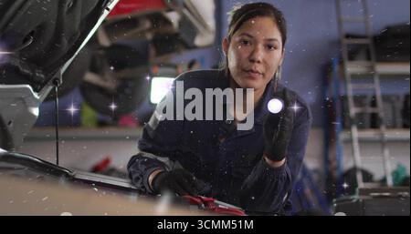 Kneeling woman mechanic in coveralls checking car engine in shop, flashlight lighting jumper clamps Stock Photo