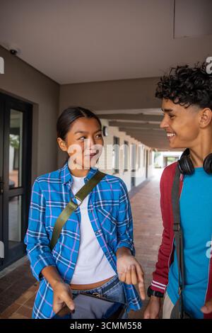 Diverse student friends walking and talking in campus corridor with tablet backpacks headphones Stock Photo
