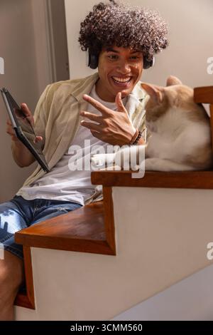 Man sitting on staircase using tablet and headphones while cat basking in sunlight Stock Photo