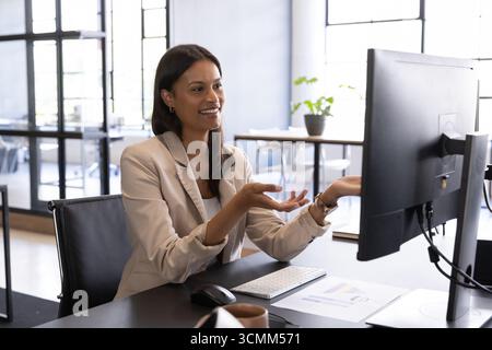 Woman in business attire gesturing toward monitor at office desk with coffee mug, charts Stock Photo