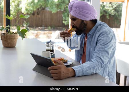 Indian man sitting at kitchen island using tablet and eating tomato toast next to coffee Stock Photo