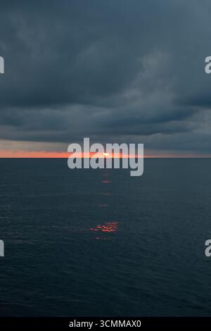 Red sunrise setting over sea under heavy, dark clouds. Moody and intense sunset scene with straight horizon line over calm sea Stock Photo