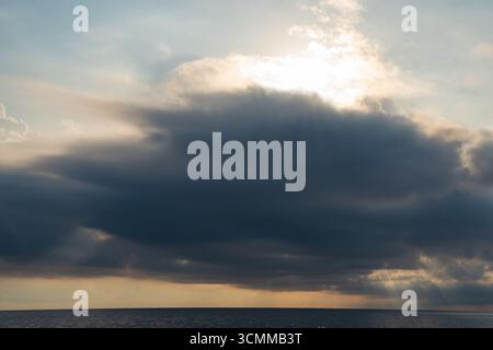 Sun rays breaking through large, dark cloud over calm ocean. Straight horizon separating calm sea from dramatic Stock Photo