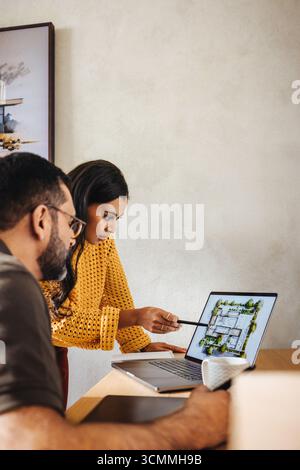 Two coworkers are engaged in a discussion over a digital architectural floor plan displayed on a laptop, emphasizing teamwork and creative design in a Stock Photo