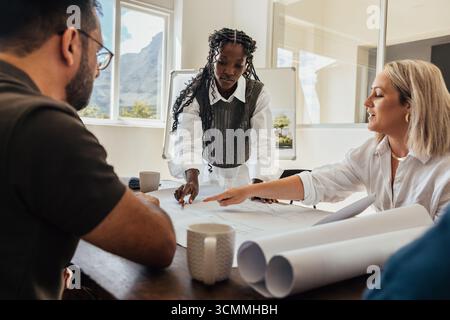 A group of coworkers, two women and a man, discussing architectural plans in a bright office. They look focused and engaged, showcasing teamwork and c Stock Photo