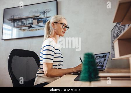 A professional mature woman architect working at a drafting table, advancing architectural designs inside a well-lit modern office. The workspace is a Stock Photo
