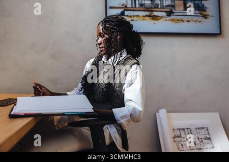 A young female architect sits at her desk with blueprints, engaged in work. The modern office environment and architectural designs emphasize creativi Stock Photo