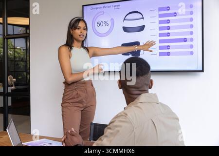 Diverse colleagues presenting charts on display, discussing data via laptop at boardroom table Stock Photo